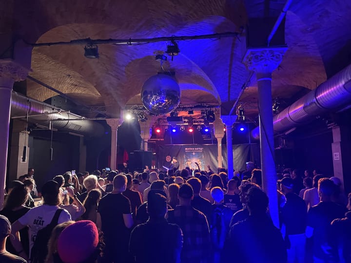 A crowd, lit by blue light stand underneath a disco ball. They look at a stage in the front of the room. 